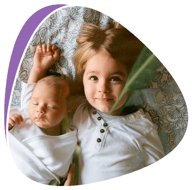 A smiling child laying beside a sleeping baby.